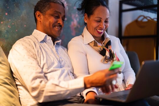 Happy adult Ethiopian married couple in elegant outfits smiling and looking at screen of laptop while entering credit card credentials into laptop at home