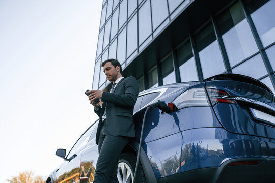 Electric car is charging, businessman with smartphone is waiting