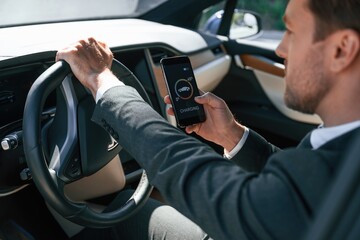 Naklejka premium Holding smartphone with charging progress app. Businessman in suit is driving his electric car