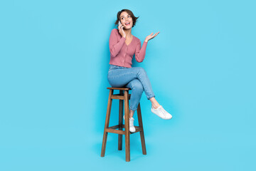 Full size photo of funny woman wear pink cardigan talk on smartphone sit on chair look empty space ad isolated on teal color background