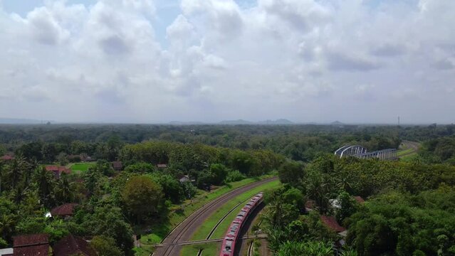 Aerial view of the  train passes through the area of green trees and and enter the metal bridge with cloudy sky during the day. Trains through the countryside, Indonesia - 4K drone footage