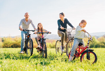 Smiling father and mother with two daughters during summer outdoor bicycle riding. They enjoy togetherness on green high grass meadow. Happy parenthood and childhood or active sport life concept image