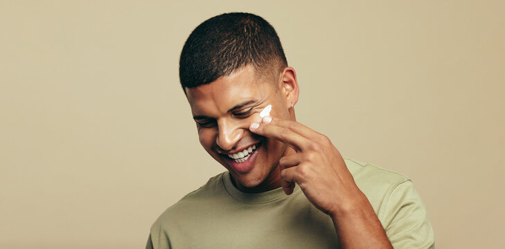 Happy with his skincare routine, young man smiles as he applies facial cream