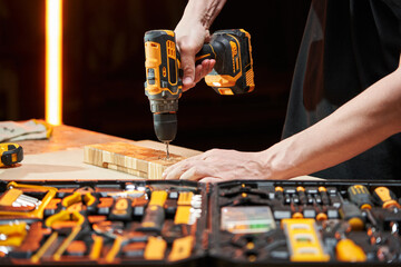 The hands of a Caucasian worker drill with an electric drill-driver with a replaceable battery a wooden part on a workbench, on which lies a case with different tools