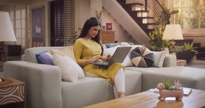 Beautiful Indian Woman Works Remotely From Home, Seated On Sofa With Laptop. Productive and Efficient at Virtual Management. Browsing Internet and doing Online Shopping. Slow Motion Arc Wide shot