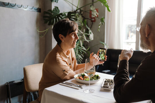 Pretty elderly woman shows photo of food on phone to her husband on date in cafe 