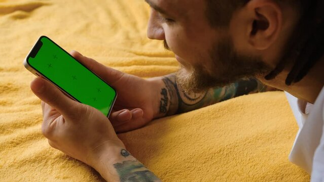 Caucasian man using smartphone in vertical mode with a green mock-up screen lies on a yellow blanket and watches videos or movies. 4k footage. Focus on Chroma Display and hands.