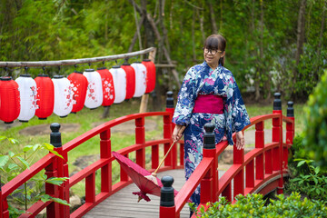 Cute Japanese women wearing beautiful dresses, casual Yukata kimonos, and red umbrellas wear traditional clothes and stand on a red wooden bridge in the garden.