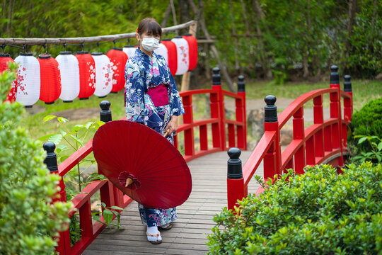 Cute Japanese women wearing beautiful traditional clothes dresses casual Yukata kimonos with white masks, and red umbrellas stand on a red wooden bridge with floral lanterns in garden.