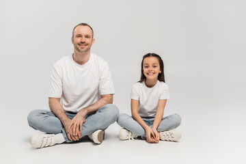 Fototapeta na wymiar happy preteen girl with long brunette hair and cheerful dad with bristle sitting with crossed legs while posing in white t-shirts and blue denim jeans on grey background, Happy Father`s Day