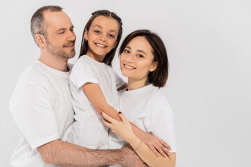 Fototapeta premium portrait of happy family in white t-shirts looking at camera and hugging each other on grey background, International child protection day, parents and daughter