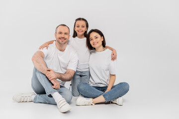 Fototapeta na wymiar stylish family in white t-shirts and blue denim jeans looking at camera and sitting together on grey background, International child protection day, preteen daughter hugging happy parents