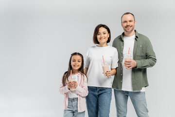 Fototapeta premium Positive parents in casual clothes looking at camera near daughter with milkshake in plastic cup during international children day celebration isolated on grey with copy space