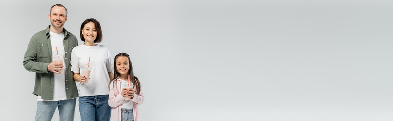 Smiling adult parents in casual clothes holding milkshakes near preteen kid and looking at camera during international child protection day isolated on grey with copy space, banner