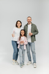 Full length of smiling family with preteen daughter in casual clothes holding milkshakes in plastic cups and looking at camera during children day celebration on grey background