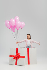 Carefree preteen kid in casual clothes looking at camera while holding pink festive balloons while standing in big present during international children day celebration on grey background