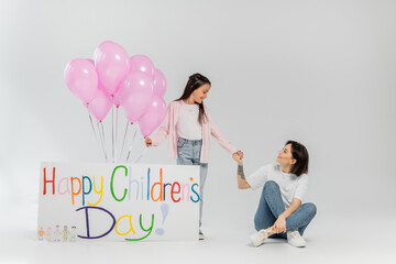 Smiling tattooed mother holding hand of preteen daughter near placard with happy children's lettering and pink festive balloons during celebration in June on grey background