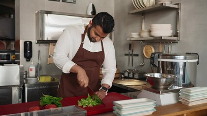 Handsome indian chef in apron slices lettuce leaves in kitchen of cafe