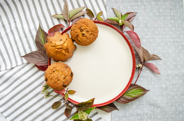 Obraz na płótnie Canvas Muffins on a ceramic plate with green and red plant leaves and wild flowers on cotton tablecloth and napkins. Mock up. Template for recipes or food menu
