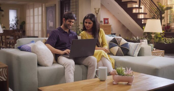 Portrait of Loving Young Indian Couple Enjoys Browsing Online Shopping Websites Together On A Laptop, While Sitting Cozily On Their Living Room Sofa. Browsing Interner, Choosing Products to Purchase