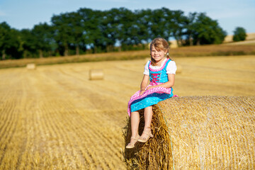 Naklejka na ściany i meble Cute little kid girl in traditional Bavarian costume in wheat field. Happy child with hay bale during Oktoberfest in Munich. Preschool girl play at hay bales during summer harvest time in Germany.
