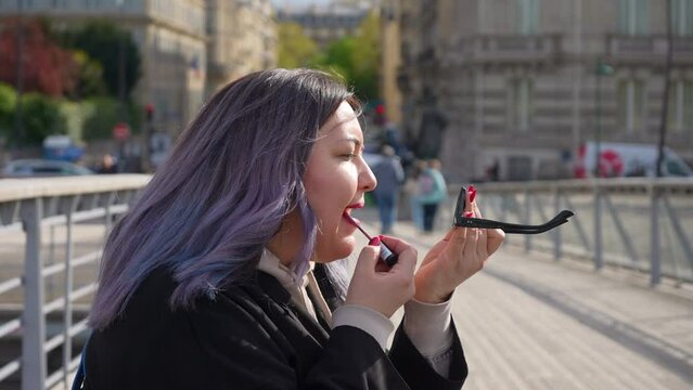 Side view of female with dyed hair applying lipstick looking at reflection in windows of sunglasses standing on city street in sunny day. Medium shot of woman applying lip pencil on lips in Paris.