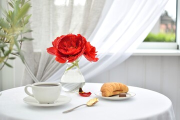 Red terry tulips in a vase on the table along with a cup of coffee and a croissant