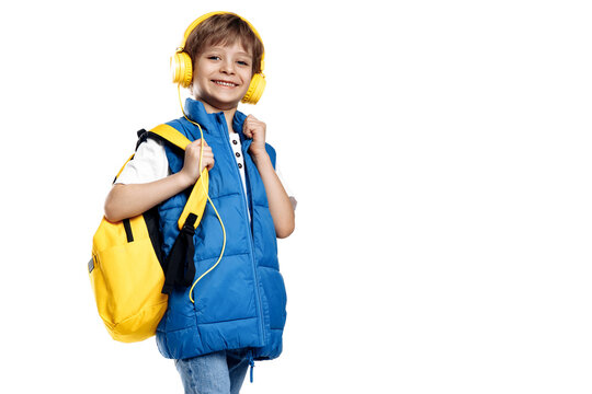 Cheerful little boy wearing headphone and yellow backpack looking to camera on white background. Back to School.