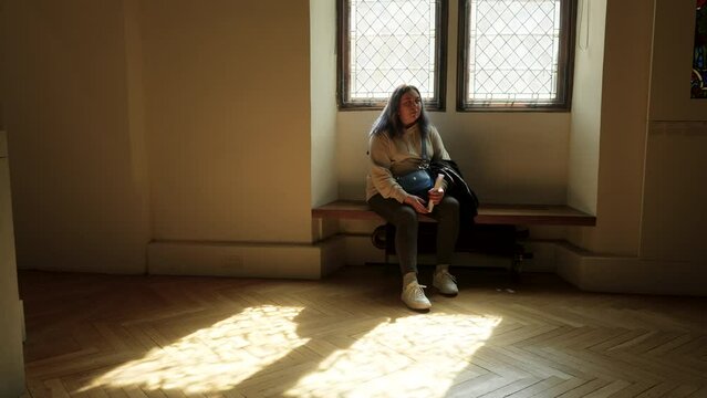 Wide shot of pensive young woman sitting on bench under windows of Paris Musee de Cluny in warm sunlight, examining exhibition. Caucasian female visits exhibition of Musee National du Moyen Age.