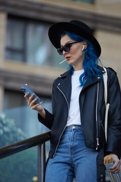 Beautiful diverse female person using a smart phone outdoor. Portrait of a stylish white woman with dyed blue hair listening to music in wireless headphones and eating a lollipop candy