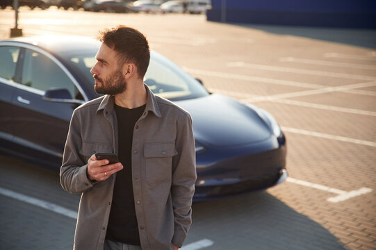 With smartphone. Man is standing near his electric car outdoors