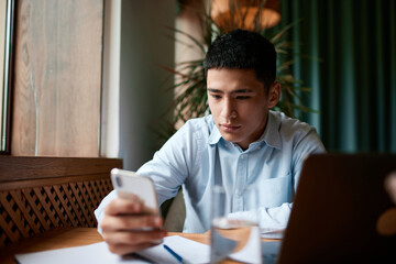 Fototapeta premium Confident busy male freelancer, businessman doing remote job using smartphone searching information for presentation in cafe