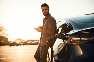 Leaning on the automobile and holding smartphone. On the charging station. Man is standing near his...