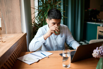 Portrait with young man, freelancer, wearing shirt sitting with laptop, overworking on project in cafe and drinking coffee. Business people