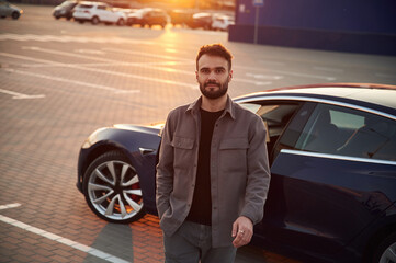 Sunny background. Man is standing near his electric car outdoors