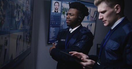 Security officer controls CCTV cameras using tablet and talks with African American coworker in surveillance center. Big screens on the wall showing security cameras footage. Social safety. Close up.