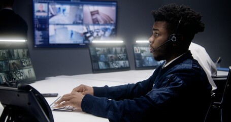 African American operator comes at workplace, puts on headset and starts controlling security cameras. Multiple big screens on the wall showing CCTV cameras footage. Colleagues working at background.