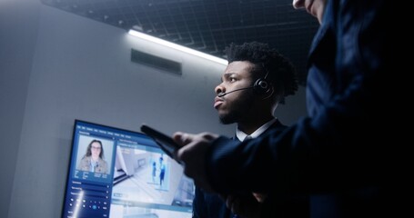 Close up of African American male officer in headset talking with colleague and monitoring CCTV...