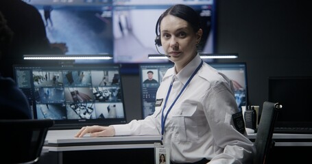 Female operator in headset in police monitoring center turns and looks at camera. Computer monitors...