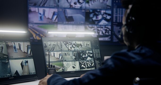 African American officer in headset controls security cameras with AI face scanning in police monitoring room. Multiple big screens with CCTV footage at background. Concept of social safety. Close up.