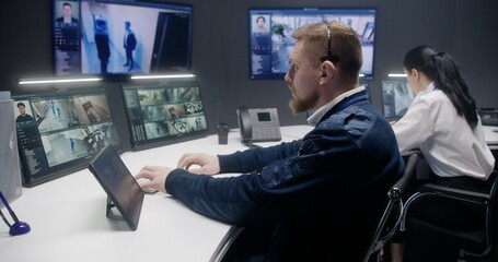 Male security officer touches tablet and controls CCTV cameras with AI face scanning. Computer monitors and big digital screens showing security cameras footage. Female colleague works at background.