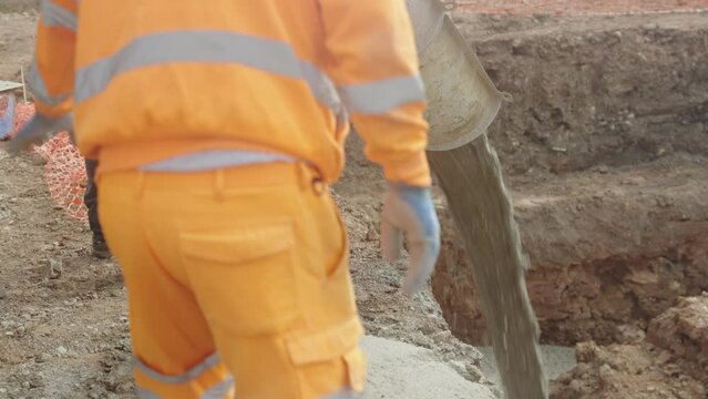 UK - cement pours into a pre-dug pit. Two workers supervise. When the cement dries, it will form the base of a house foundation.