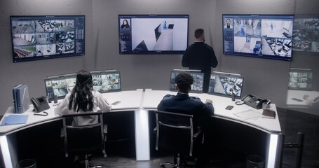 Multi ethnic security officers monitor CCTV cameras with AI face scanning. Multiple big screens with displayed security cameras. Teamwork in surveillance room. Monitoring, tracking and social safety.