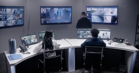 Multi ethnic security officers monitor CCTV cameras with AI face scanning. Multiple big screens with displayed security cameras. Teamwork in surveillance room. Monitoring, tracking and social safety.