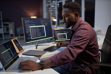 African American programmer concentrating on his online work on computers while working in office till late evening - obrazy, fototapety, plakaty