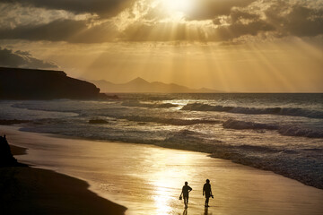 Fototapeta na wymiar Sonnenuntergang am Strand von La Pared, Fuerteventura - ein Mann und eine Frau stehen allein im beleuchteten nassen Sand, ihre Schuhe in der Hand