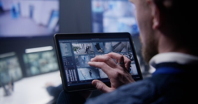 Close up shot of security officer zooming CCTV cameras with AI facial scanning using tablet in police surveillance center. Colleagues work at background. Monitoring system. Concept of social safety.