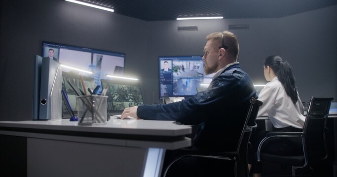 Male security operator in headset controls CCTV cameras with AI facial recognition in police monitoring center. Tablet, PC monitors and big digital screens showing security cameras footage. Tracking.