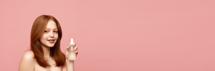 Young, beautiful, redhead girl with healthy, well-kept skin holding face toner against pink...