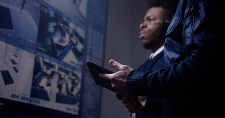 Security officer controls CCTV cameras using tablet and talks with African American coworker in surveillance center. Big screens on the wall showing security cameras footage. Social safety. Close up.